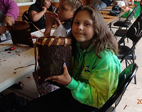 A girl showing off her cedar basket along with other children who are working on making them